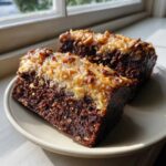 Two moist slices of german chocolate poke cake with a thick, gooey coconut-pecan topping sitting on a plate near a window.