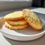 A stack of three golden brown ultimate sugar cookie rounds served on a white plate, with one leaning against the stack.