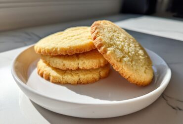 A stack of three golden brown ultimate sugar cookie rounds served on a white plate, with one leaning against the stack.