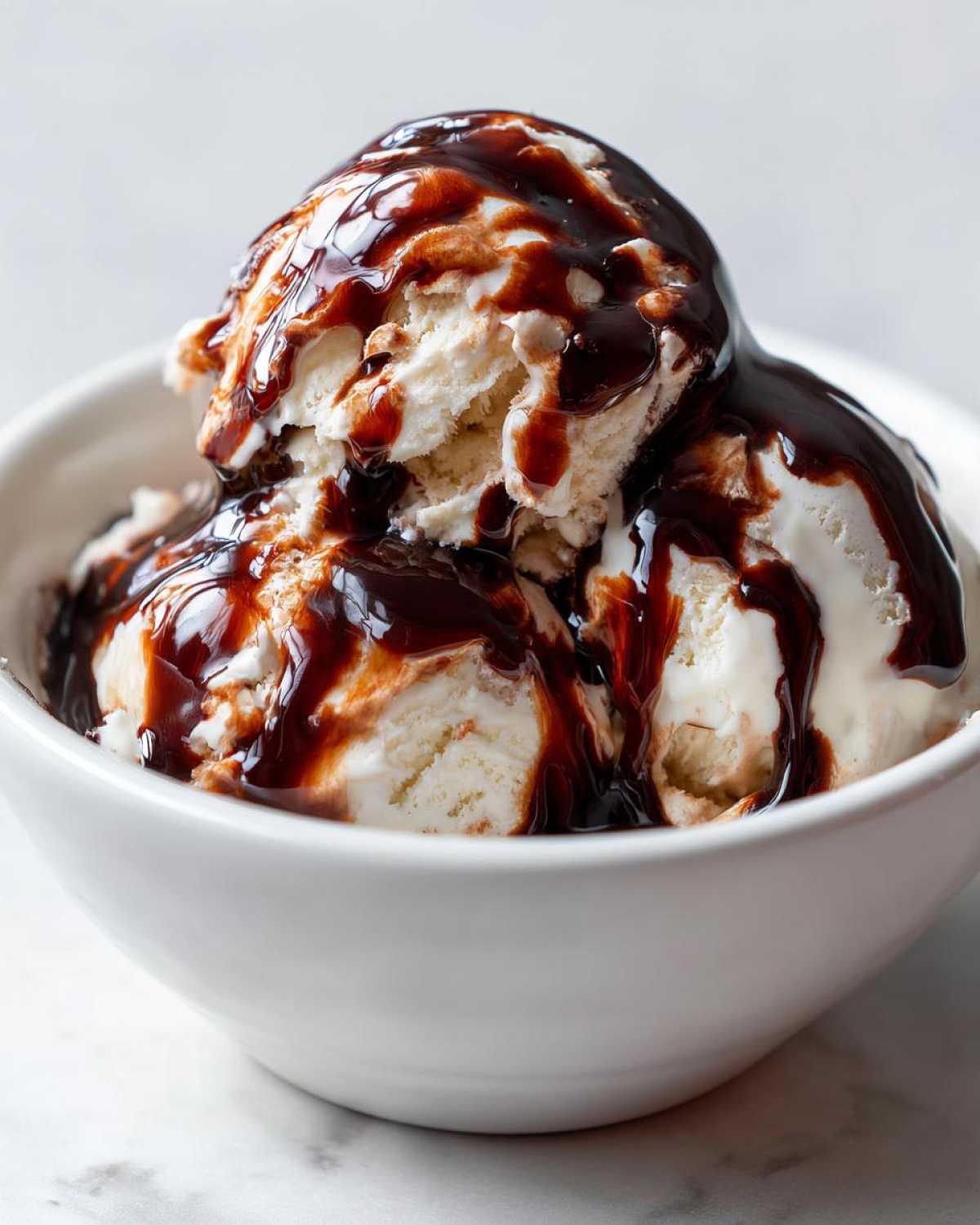 Two scoops of vanilla ice cream generously covered in homemade chocolate syrup in a white bowl.