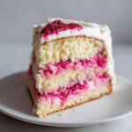 A tall slice of white chocolate raspberry cake showing three layers of vanilla sponge and bright pink raspberry filling.