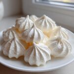 A close-up of several perfectly piped, white meringue cookies stacked on a small white plate near a window.