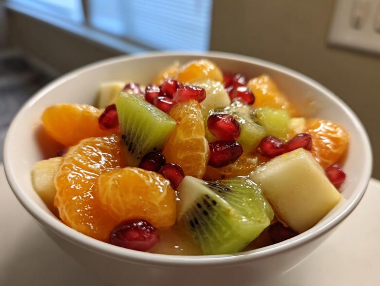 Close-up of a vibrant winter fruit salad featuring mandarin oranges, kiwi slices, and pomegranate seeds in a white bowl.
