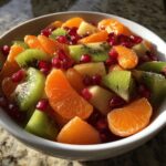 Close-up of a vibrant winter fruit salad featuring mandarin oranges, kiwi slices, and pomegranate seeds in a white bowl.