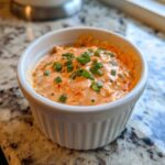 Close-up of zesty remoulade sauce, orange in color and topped with fresh green chives, served in a white ramekin.