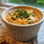 A close-up of zesty remoulade sauce, orange-hued and topped with fresh green chives, in a white ramekin.