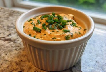 A close-up of zesty remoulade sauce, orange-hued and topped with fresh green chives, in a white ramekin.