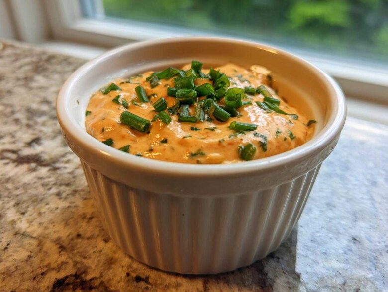 A close-up of zesty remoulade sauce, orange-hued and topped with fresh green chives, in a white ramekin.