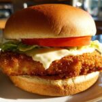 Close-up of an Air Fryer Crispy Fish Sandwich featuring a golden breaded fillet, melted cheese, lettuce, and tomato on a bun.