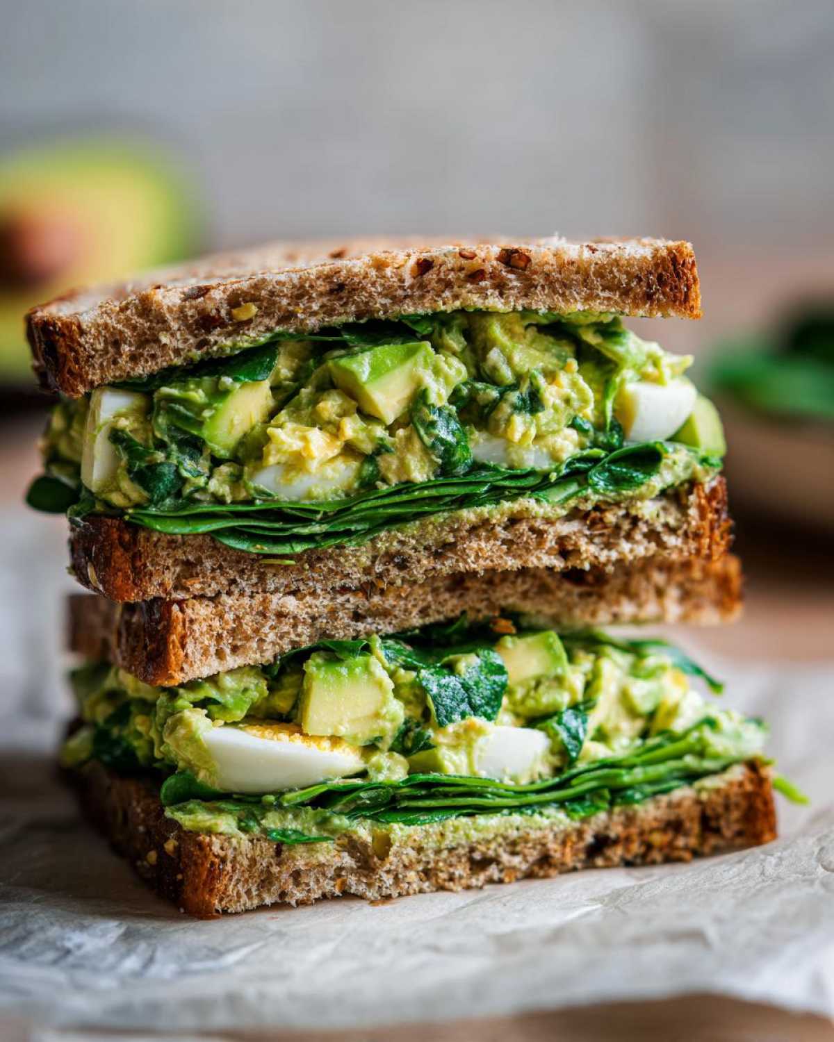 A tall, stacked Avocado Egg Salad Sandwich made with whole wheat bread, spinach, and chunks of avocado.
