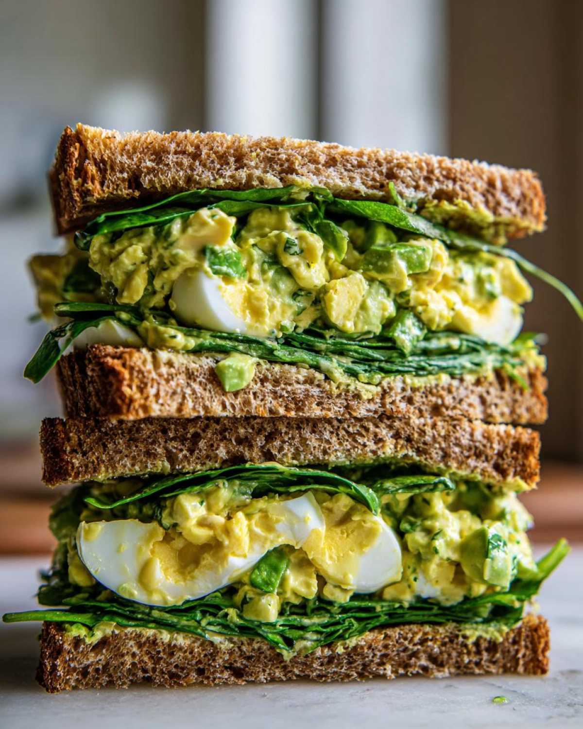 A tall, stacked Avocado Egg Salad Sandwich featuring creamy avocado filling, sliced hard-boiled eggs, and spinach on whole wheat bread.