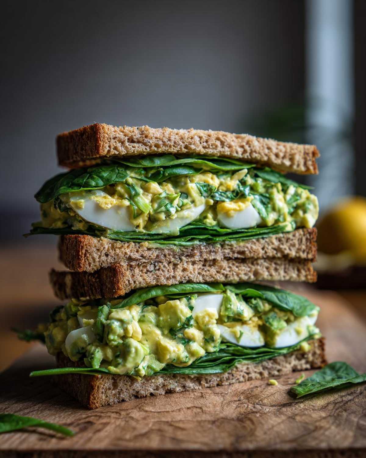Two halves of a stacked Avocado Egg Salad Sandwich featuring creamy filling, sliced hard-boiled eggs, and fresh spinach on whole wheat bread.