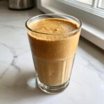 A tall glass filled with a thick, creamy Banana Peanut Butter Smoothie sitting on a white marble countertop.