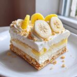 A square slice of Banana Pudding Cheesecake Bars with layers of crust, cream, and banana pudding, topped with whipped cream and fresh banana slices.