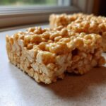 Two thick, chewy squares of Brown Butter Rice Krispie Treat Bars sitting on a light countertop near a window.