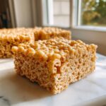 A close-up, appetizing shot of a freshly cut Brown Butter Rice Krispie Treat Bar sitting on a white marble surface.