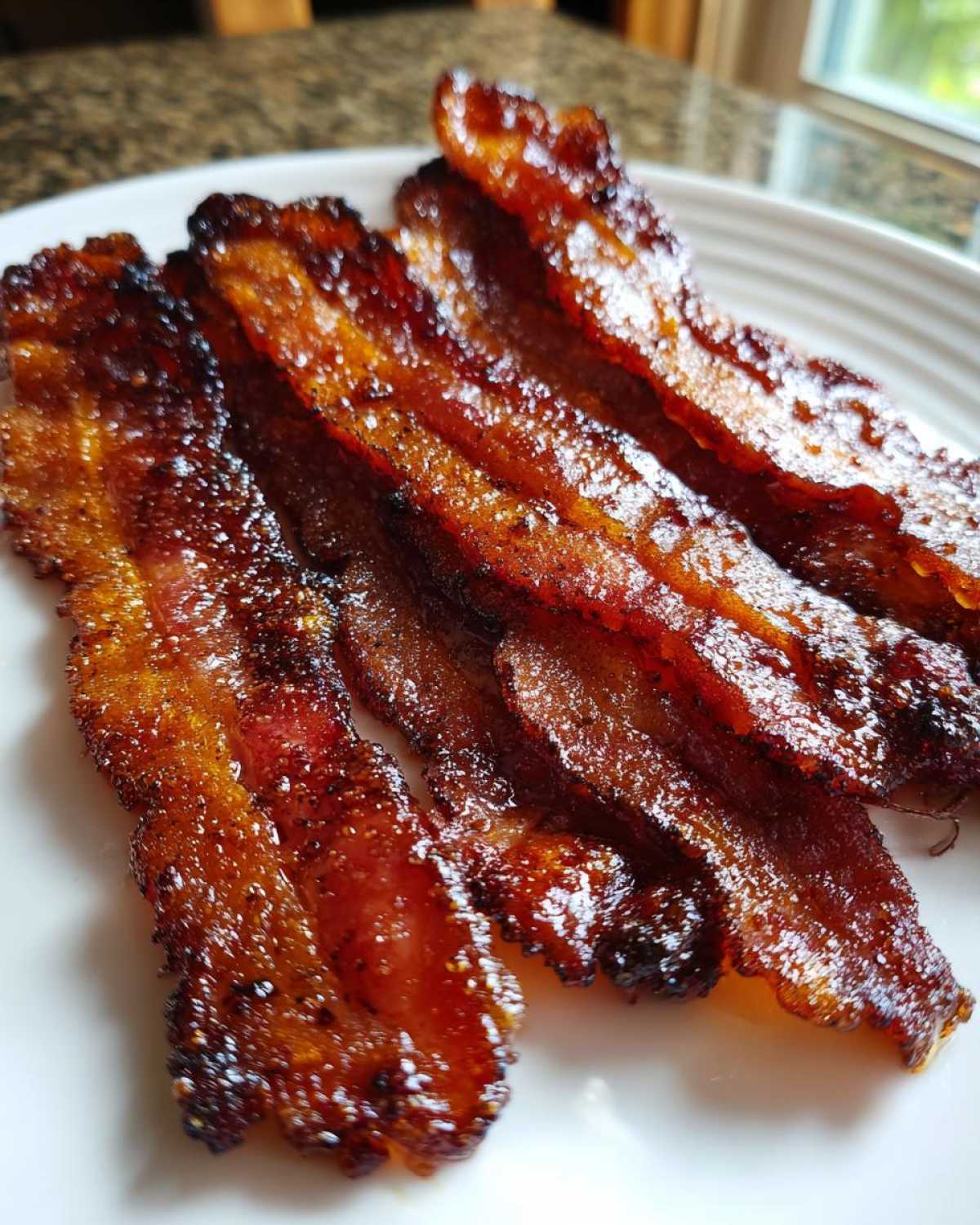 Four strips of crispy, shiny Brown Sugar Glazed Bacon resting on a white plate.