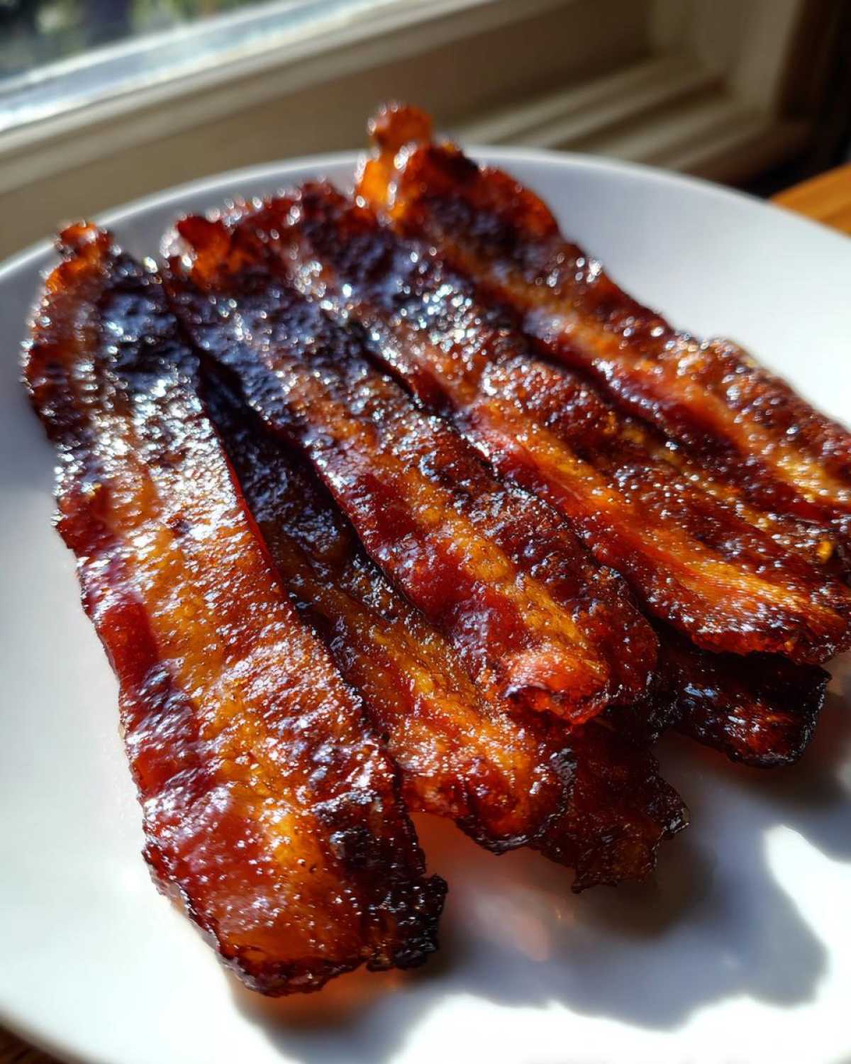 Close-up of four strips of shiny, caramelized Brown Sugar Glazed Bacon resting on a white plate near a window.