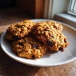 Four chewy Gluten-Free Oatmeal Raisin Cookies piled on a white plate, illuminated by natural light.