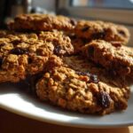 Close-up of several chewy Gluten-Free Oatmeal Raisin Cookies piled on a white plate, illuminated by sunlight.