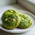 Three vibrant green, chewy matcha white chocolate cookies resting on a white plate near a window.