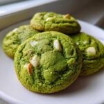 A close-up of four vibrant green Chewy Matcha White Chocolate Cookies studded with white chocolate chunks, resting on a white plate.