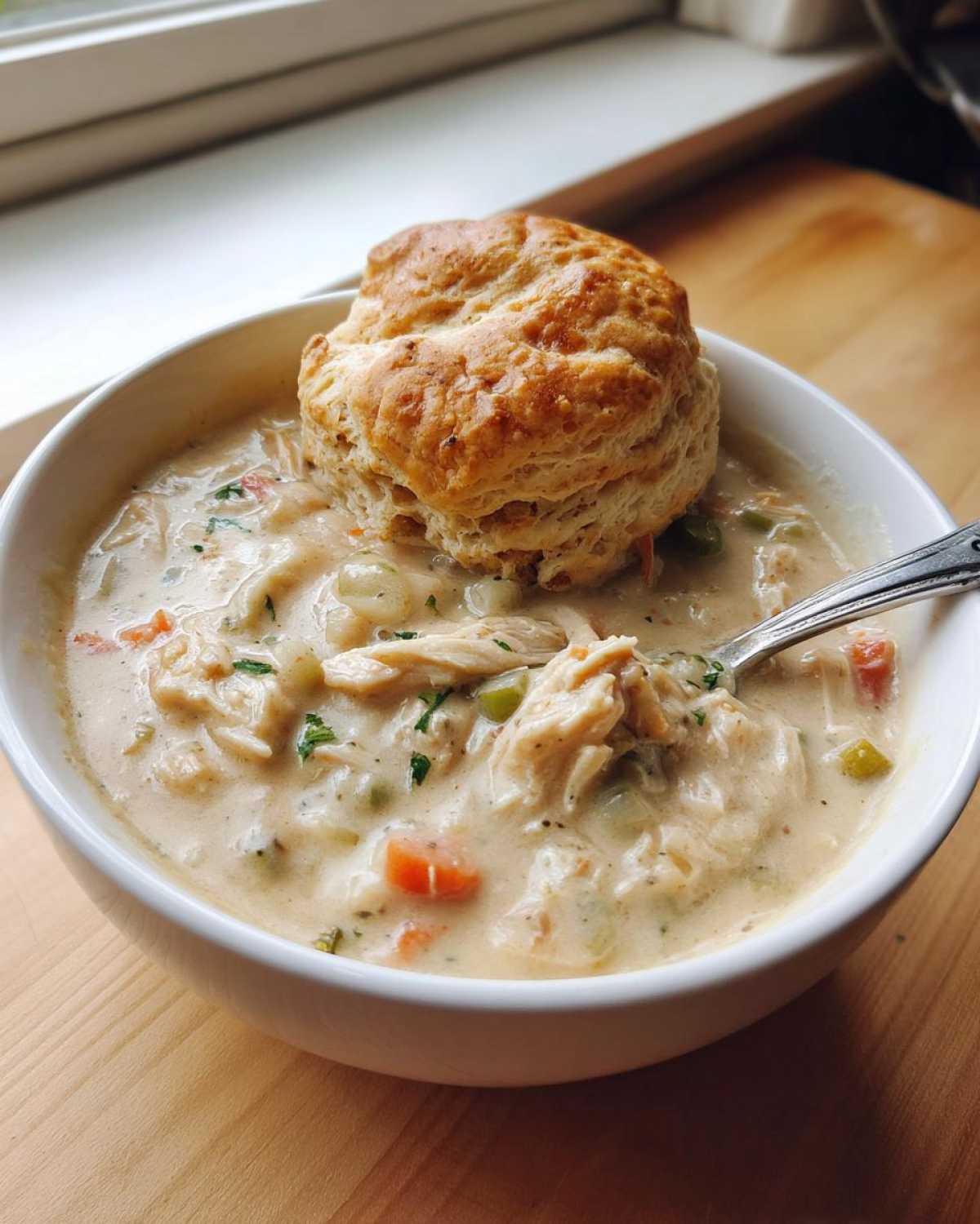 A bowl of creamy Chicken Pot Pie Soup topped with a golden biscuit, ready to eat with a spoon.