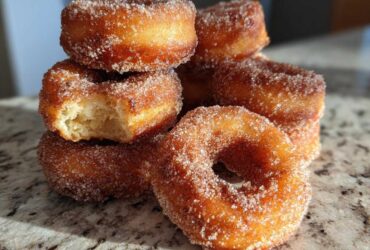 A stack of golden brown Cinnamon Sugar Air Fryer Donuts heavily coated in sparkling cinnamon sugar, one has a bite missing.