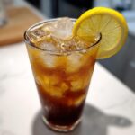 A tall glass filled with iced Cold Brew Lemonade, showing condensation and garnished with a bright lemon slice.