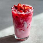 A tall glass filled with the Copycat Starbucks Pink Drink, showing swirling pink and white layers and topped with fresh sliced strawberries.