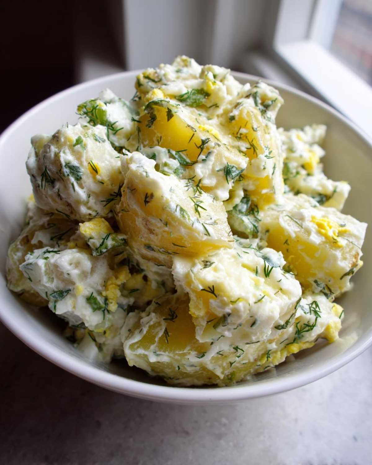 Close-up of chunky potatoes coated in creamy dressing, fresh dill, and egg pieces in a white bowl, featuring Creamy Dill Potato Salad.