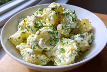 A close-up of a white bowl filled with chunks of potato coated in a creamy dressing and generously topped with fresh dill for Creamy Dill Potato Salad.