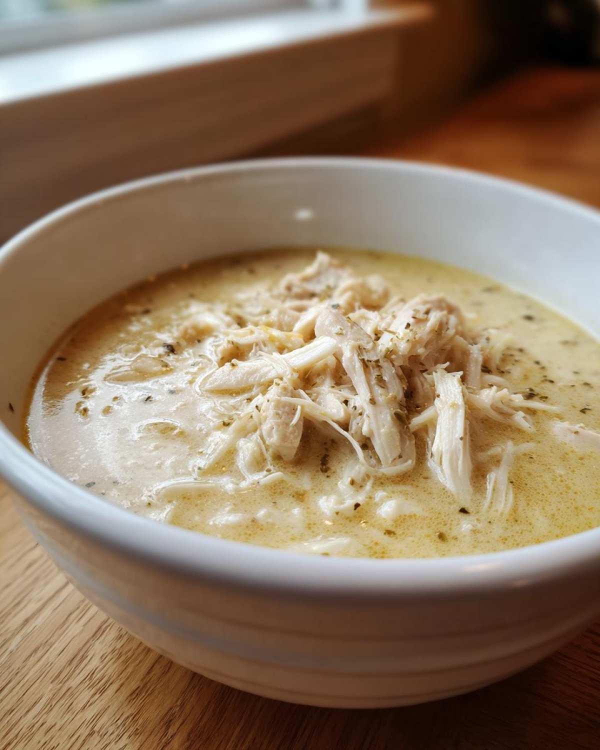 Close-up of a white bowl filled with creamy Greek Lemon Chicken Soup, topped generously with shredded chicken and herbs.