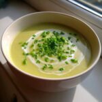 A close-up of a bowl of vibrant Creamy Potato Leek Soup, garnished with a swirl of cream and fresh chopped chives.