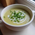 A white bowl filled with vibrant Creamy Potato Leek Soup, garnished with a swirl of cream and fresh chopped chives.