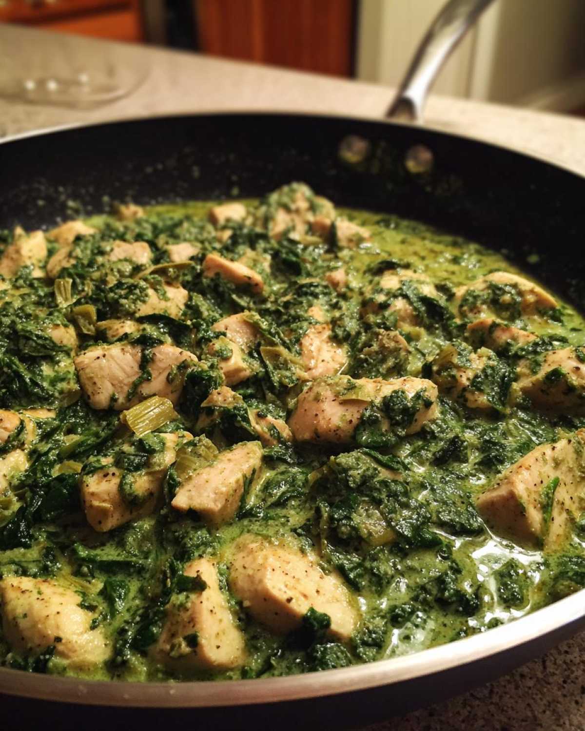 Close-up of cubed chicken pieces simmering in a vibrant green, creamy spinach artichoke sauce in a black skillet.