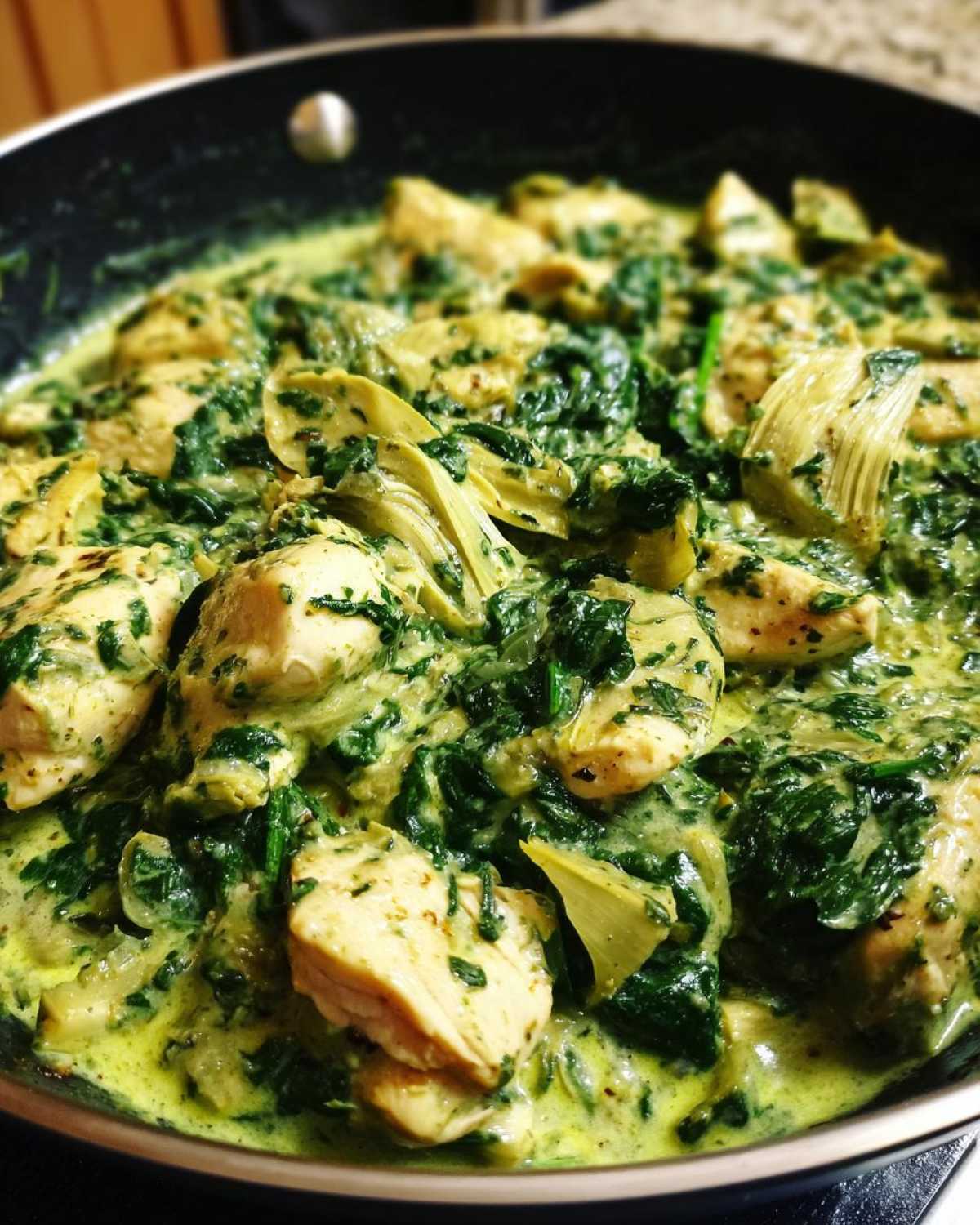 Close-up of Creamy Spinach Artichoke Chicken Skillet cooking in a black pan with chunks of chicken.