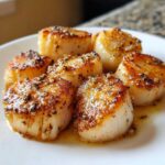 Close-up of six golden-brown Garlic Butter Scallops with Lemon sauce and cracked black pepper on a white plate.