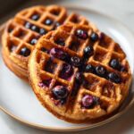 Close-up of two golden brown Gluten-Free Blueberry Waffles topped with juicy, burst blueberries.
