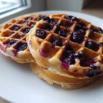 Close-up of two golden Gluten-Free Blueberry Waffles stacked on a white plate near a window.