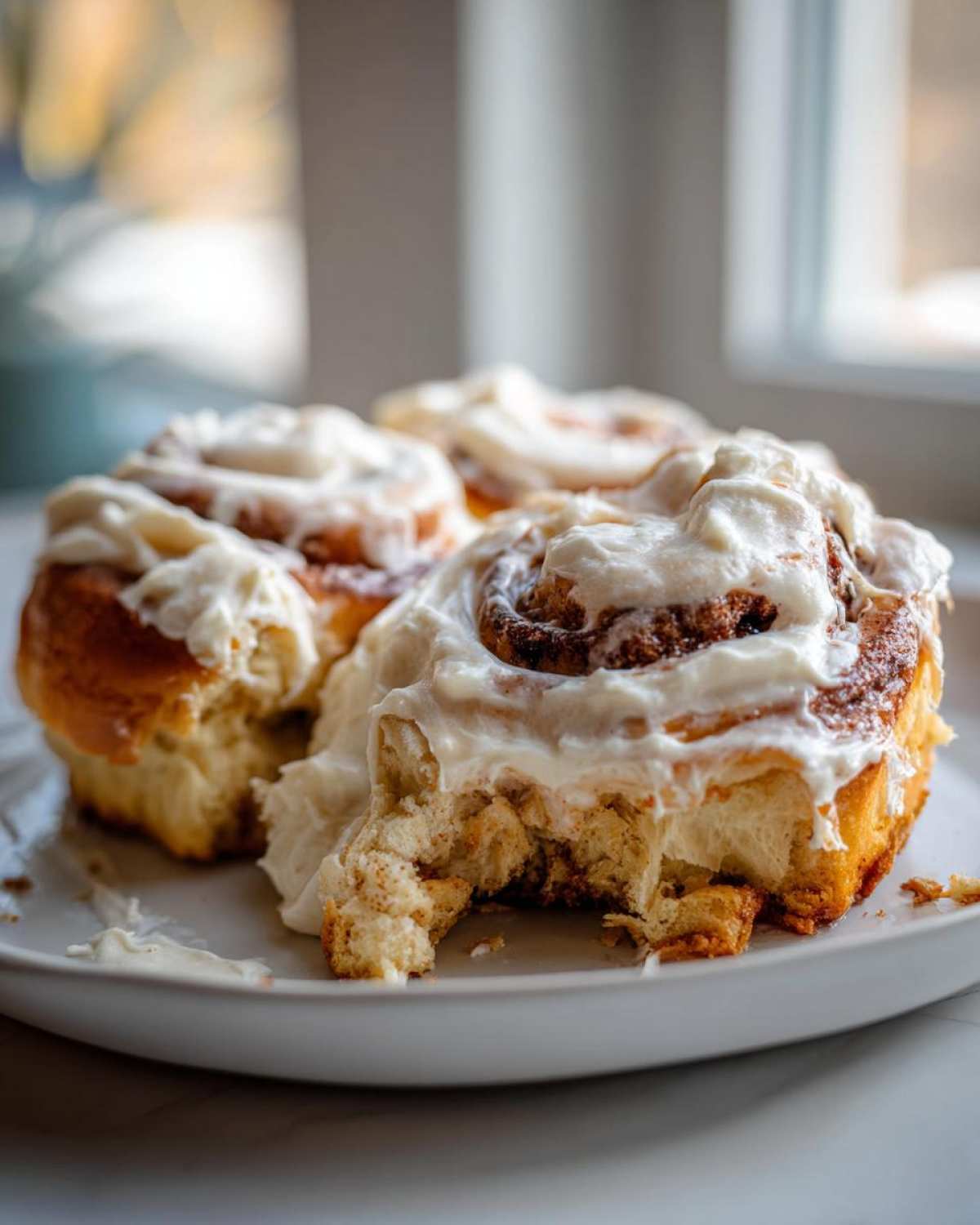 Close-up of three soft Gluten-Free Cinnamon Rolls piled on a plate, heavily topped with cream cheese frosting; one roll has a bite taken out.