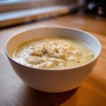A close-up of a steaming white bowl filled with creamy Greek Lemon Chicken Soup, topped generously with shredded chicken and orzo.