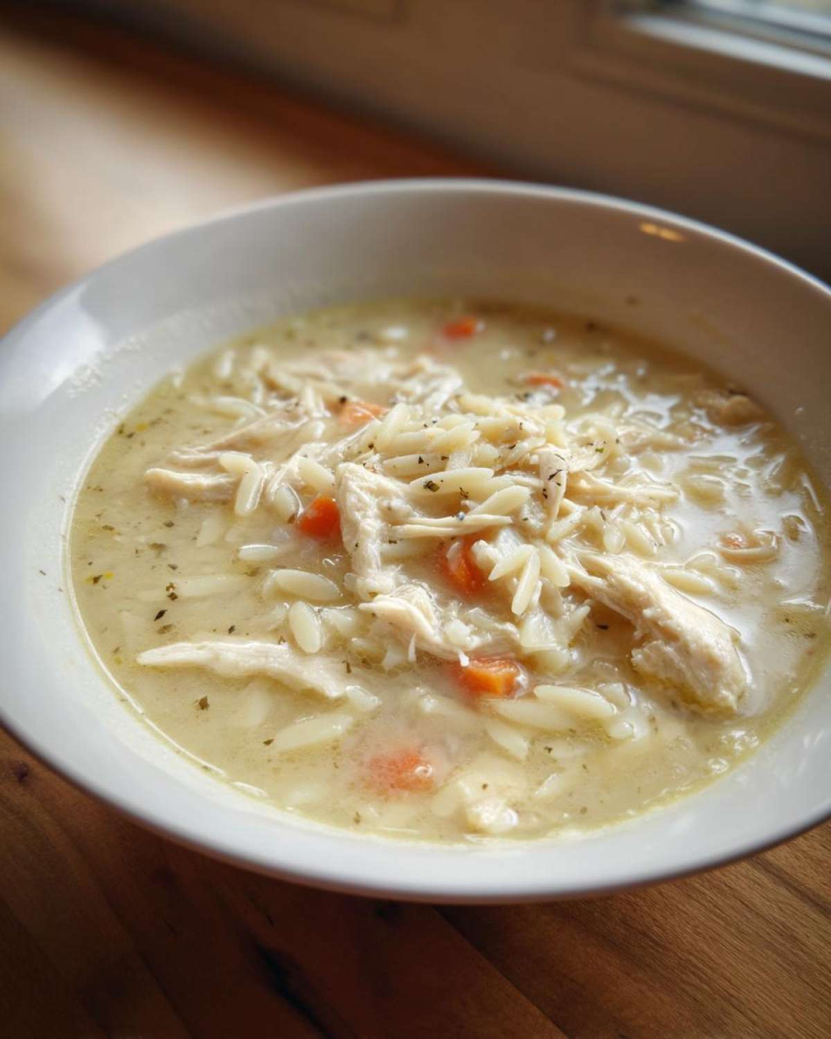 Close-up of a white bowl filled with creamy Greek Lemon Chicken Soup, featuring shredded chicken, orzo pasta, and carrots.