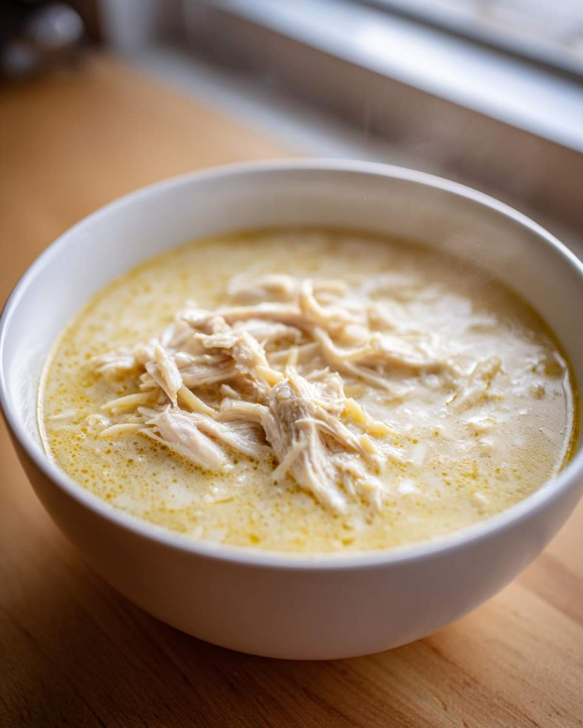 Close-up of a white bowl filled with steaming Greek Lemon Chicken Soup topped with shredded chicken.