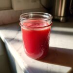 A glass mason jar filled with vibrant red Homemade Strawberry Lemonade Concentrate sitting on a sunlit marble counter.