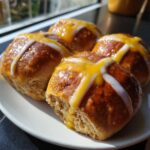 Four freshly baked Hot Cross Buns with Orange Glaze, drizzled with white icing, sitting on a white plate.