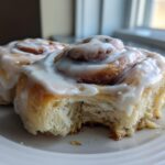 Two fluffy Gluten-Free Cinnamon Rolls covered in thick white icing, one with a bite taken out.