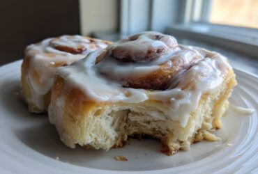 Two fluffy Gluten-Free Cinnamon Rolls covered in thick white icing, one with a bite taken out.
