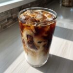 A tall glass of Iced Honey Almond Milk Latte showing rich swirls of coffee mixing with white almond milk and ice cubes.