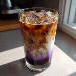 Close-up of a layered Iced Lavender Latte with ice, purple syrup, milk swirling into coffee, topped with lavender buds.